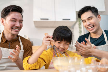 Asian attractive LGBTQ gay family teach girl kid making yeast dough. Handsome male couple take care and spend time with little adorable child bake bakery in kitchen, enjoy parenting activity at home.の写真素材