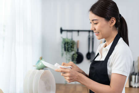 Asian active cleaning service woman worker cleaning in kitchen at home. Beautiful young girl housekeeper cleaner feel happy and wipes dishes plates after washing for housekeeping housework or chores.の写真素材