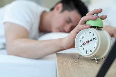 Asian attractive young man turn off alarm clock ringing in the morning. Handsome male feel lazy and...