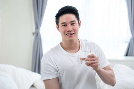 Portrait of Asian active strong man holding clean water in bedroom. Attractive thirsty male drink or take a sip of mineral natural in cup after wake up for health and wellbeing and looking at camera.の写真素材