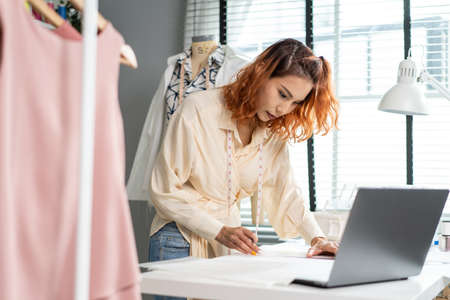 Asian beautiful tailor woman working on clothes in tailoring atelier. Attractive female fashion designer dressmaker design drawing and sketching out ideas for new fashion collection in workshop room.の写真素材