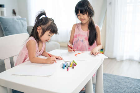Asian young girl children drawing and coloring paper in living room. Little adorable sibling child sister sit on table, learn how to sketch art picture enjoy creativity activity on holiday at home.の写真素材