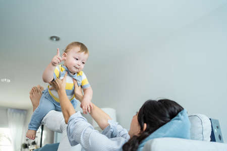 Caucasian loving mom play with baby boy child on sofa in living room. Happy family, attractive beautiful caring young mother lifting toddler son fly up enjoy doing airplane game in morning in house.の写真素材