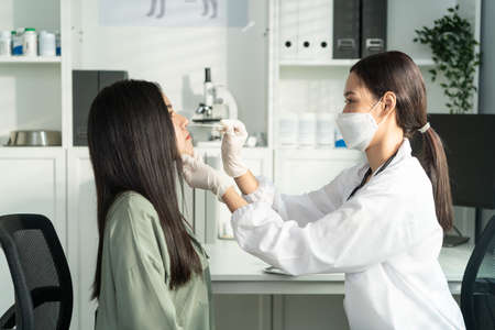 Asian young woman doctor using flashlight on patient's eye in hospital. Attractive specialist physician wear mask, examining and checking eyesight for lady in clinic. Medical insurance service conceptの写真素材