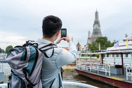 Asian young man backpacker using mobile phone take a picture in city. Handsome male tourist traveler travel on street use cellphone record video on holiday vacation trip in Buddha temple in Thailand.の写真素材