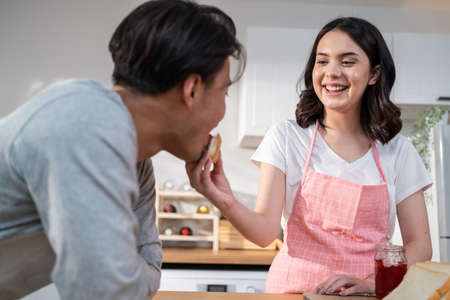 Asian young new marriage couple spend time together in kitchen at home. Happy family, Attractive beautiful women enjoy cook food for breakfast, feed bread into boyfriends' mouth with happiness at homeの写真素材