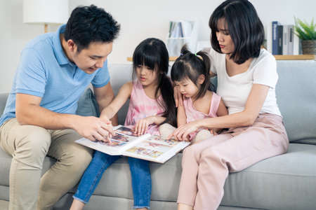 Asian family look at photographs on old picture album recalls the past. Young beautiful couple sitting with little kid daughters flipping pages of photo book and enjoy remembering relationship memory.の写真素材