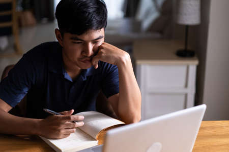 Asian young frustrated homeschool man feel bore while study at home. Attractive handsome male learn virtual internet online class from school teacher by remote meeting due to covid19 pandemic in houseの写真素材