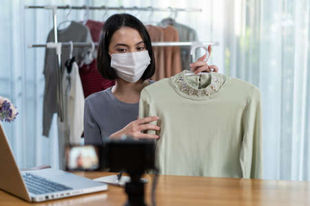 Asian attractive telesale girl selling product online live streaming. Young woman wear mask showing cloth to customer and present detail. Remote buying and purchasing technology during Covid pandemic.の写真素材