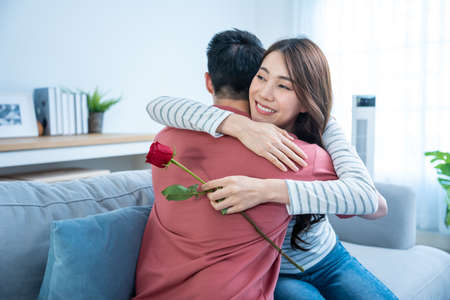 Asian young man surprise and give rose flower to beautiful girlfriend. Attractive romantic new marriage couple male and woman spend time to celebrate anniversary and valentine's day together in house.の写真素材