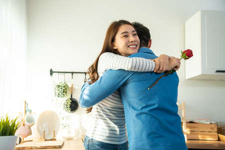 Asian young man surprise and give rose flower to beautiful girlfriend. Attractive romantic new marriage couple male and woman spend time to celebrate anniversary and valentine's day together in house.の写真素材