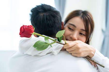 Asian young man surprise and give rose flower to beautiful girlfriend. Attractive romantic new marriage couple male and woman spend time to celebrate anniversary and valentine's day together in house.の写真素材
