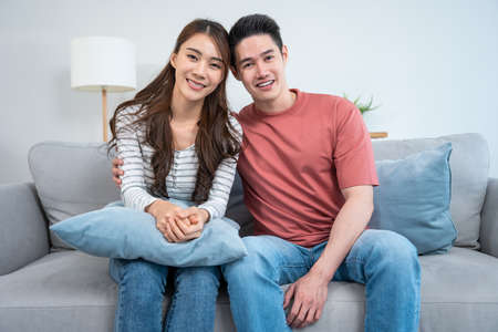Portrait of Asian young loving couple sitting on sofa in living room. Attractive romantic new marriage man and woman smiling and looking at camera enjoy spend time together on valentines day at home.の写真素材