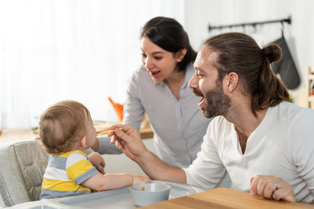 Caucasian beautiful parents take care of baby boy toddler in kitchen. Happy family, Attractive young couple cook and feed healthy foods to little kid son infant while sit on chair for lunch in house.の写真素材