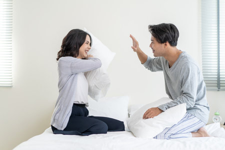 Asian young beautiful loving couple have pillow fight together on bed. Attractive new marriage man and woman feel happy, enjoy early morning activity playing in bedroom in house. Family relationship.の写真素材