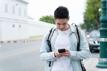 Asian attractive male backpacker chatting on mobile phone walk in city. Active handsome man tourist traveler travel alone on street use smartphone enjoy spend time on holiday vacation trip in Thailandの写真素材