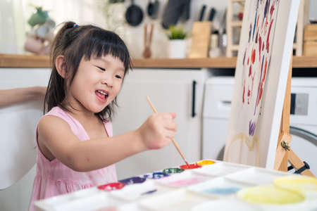 Asian young happy kid girl coloring on painting board in living room. Little adorable child learn how to draw art picture with watercolor paint and brush enjoy creativity activity on holiday at home.の写真素材