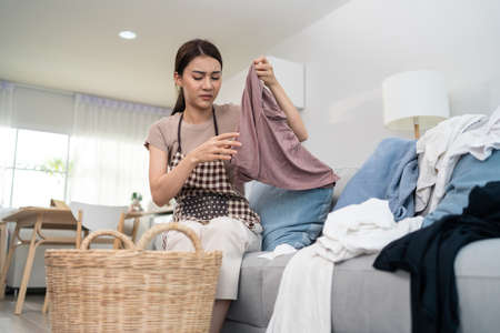 Asian cleaning service woman worker cleaning in living room at home. Beautiful young girl housekeeper cleaner feel frustrated to take messy dirty cloth into basket for housekeeping housework in house.の写真素材