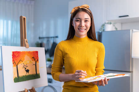 Portrait of Asian young artist girl coloring on painting board at home. Attractive female drawing art picture, creating artwork with watercolor and brush enjoy creativity activity and look at camera.の写真素材