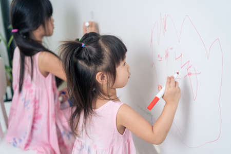Asian young sibling kid girl enjoy paint on white wall in living room. Little adorable children having fun drawing and coloring art picture with hapiness enjoy creativity activity on holiday at home.の写真素材