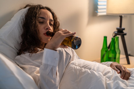 Drunk Latino woman hold beer bottle, and crying feel hangover on bed. Young beautiful depression female lying and crying in tears in bedroom, feel heart broken and celebrate life with alcohol at nightの写真素材