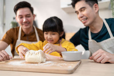 Asian attractive LGBTQ gay family teach girl kid making yeast dough. Handsome male couple take care and spend time with little adorable child bake bakery in kitchen, enjoy parenting activity at home.の写真素材