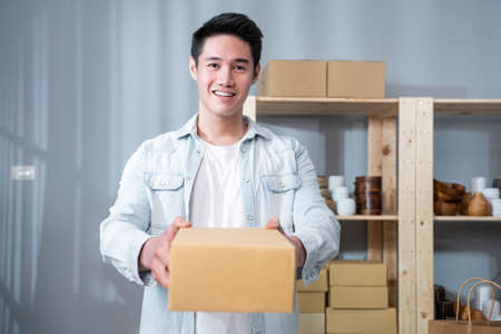 Portrait of Asian young businessman handing product order to customer. Attractive handsome delivery male smile and send shopping bag to female's hand for shipping online retail to sell at home store.の写真素材