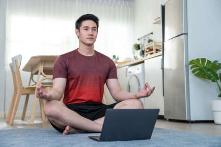 Asian handsome active young man doing exercise on floor in living room. Attractive male in sportswear spend leisure activity time to workout for health by follow online instruction on laptop at home.の写真素材