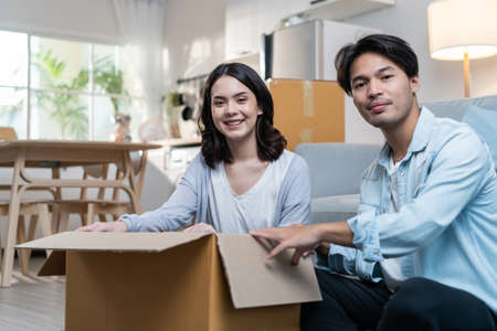 Portrait of Asian young new marriage couple move to new house together. Attractive romantic man and woman packing box parcel and suitcase and looking at camera. Family-Moving home relocation concept.の写真素材