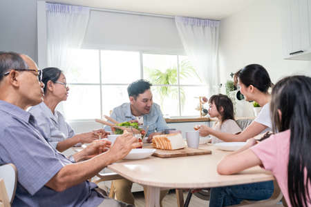 Asian big happy family have lunch on eating table together in house. Senior elder grandparent, young couple and little kid daughters feel happy, enjoy eat bread in house. Activity relationship conceptの写真素材