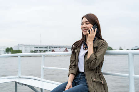 Asian young woman backpacker talk on mobile phone while sit on ferry. Attractive beautiful girl...