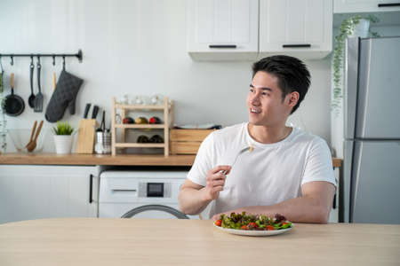 Asian attractive handsome male eating green salad in kitchen at home. Active young man sit on table,...