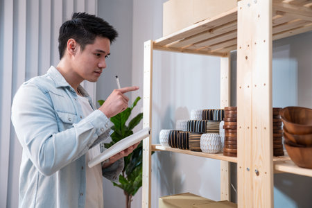 Asian business male checking vase goods order for customer from shelf. Young attractive handsome man working to preparing parcel boxes checking ecommerce shipping online retail to sell at home store.の写真素材