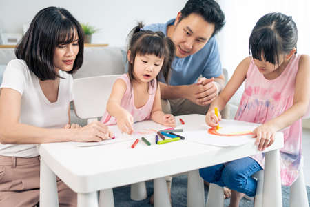 Asian happy family young couple teach little girl paint in living room. Adorable daughter feel enjoy and fun to spend activity leisure time coloring or drawing on paper with mother and father at home.の写真素材