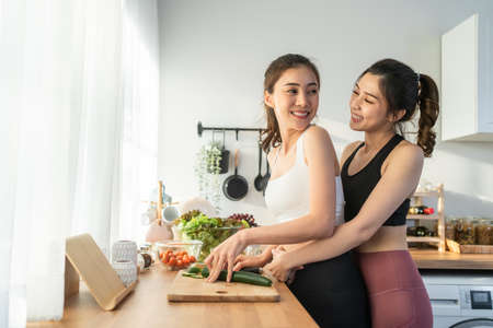 Asian young two women couple in sportswear cooking salad in kitchen. Active beautiful female hugging girlfriend and feel enjoy making vegetables healthy foods to diet and lose weight for health care.の写真素材