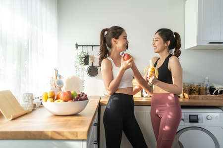 Asian active two women sibling in sportswear eat an apple in kitchen. Young beautiful girl sister feeling happy and enjoy eating fruits healthy foods to diet and lose weight for health care in house.の写真素材
