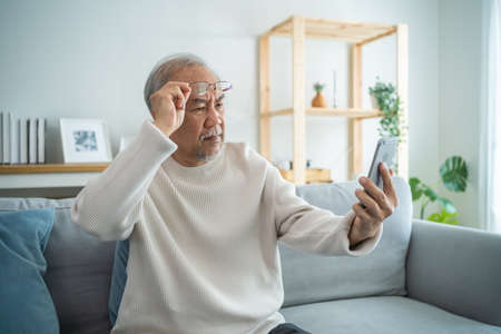 Asian senior elderly male using mobile phone in living room at home. Strong older mature grandfather...