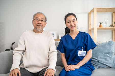 Portrait of Asian caregiver nurse sit on sofa and take care senior man. Attractive specialist carer women support older elderly mature grandfather at nursing home care and smiling, looking at camera.の写真素材