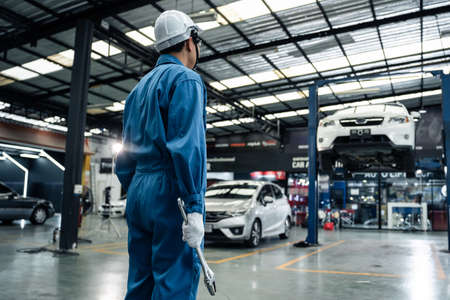 Rear view of Asian automotive mechanic repairman look under car condition in garage. Vehicle service male work in mechanics workshop, feel success after check andmaintenance to repair car engine car.の写真素材
