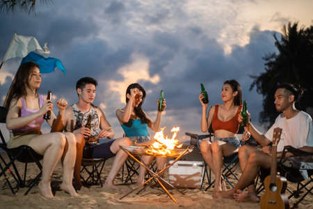 Group of Asian young man and woman having party on the beach at night. Attractive friends traveler sing and dancing while camping at seaside enjoy holiday vacation trip in tropical sea island togetherの写真素材