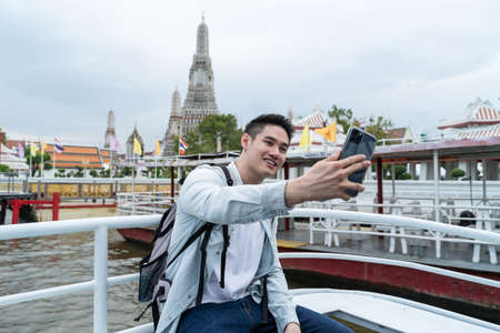 Asian young male backpacker use smartphone selfie take picture in city. Attractive handsome man tourist traveler travel alone on street, use smartphone record vlog on holiday vacation trip in Thailandの写真素材