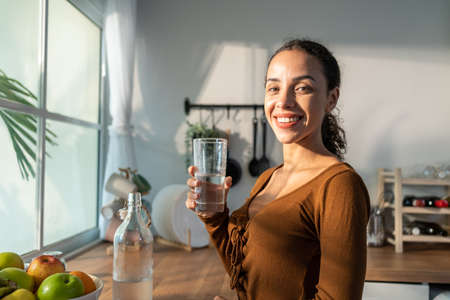 Young beautiful Latino woman holding clean water into glass in kitchen. Attractive active thirsty girl drink or take a sips of mineral natural in cup for health care and wellbeing in kitchen in house.の写真素材