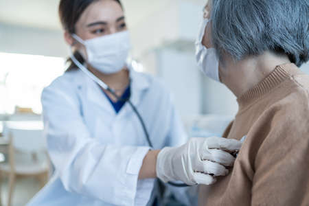 Asian nurse wear mask, take care of senior elder woman at nursing home. Beautiful therapist doctor measure heart rate by stethoscope on female older patient on sofa. Medical insurance service concept.の写真素材