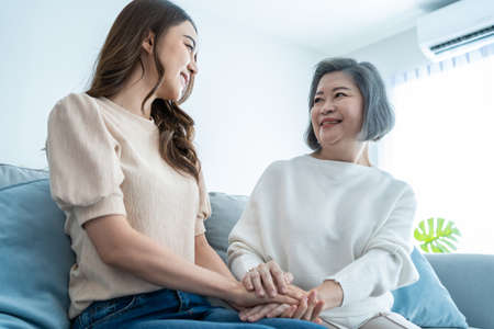 Asian lovely family, young daughter hugging and sit with older mother. Attractive woman granddaughter visit senior elder mature mom in house enjoy spend leisure time together in living room and smile.の写真素材