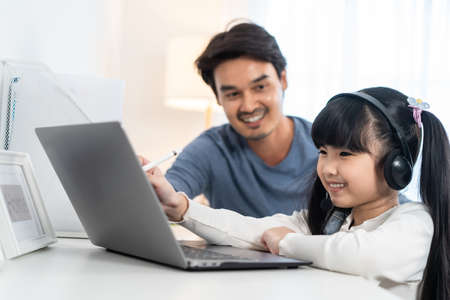 Asian little young girl kid learning online class at home with father. Preschool child use laptop...