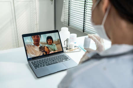 Asian veterinarian girl talk to pet owner on online virtual video call. Professional vet doctor woman use laptop computer to give treatment plan consult for little cat animal while sit in pet hospitalの写真素材