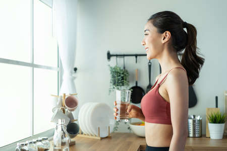 Asian beautiful woman in sportswear drink water after exercise at home. Young thirsty active sport girl takes a sips of clean mineral natural in cup after workout for health care in kitchen in house.の写真素材