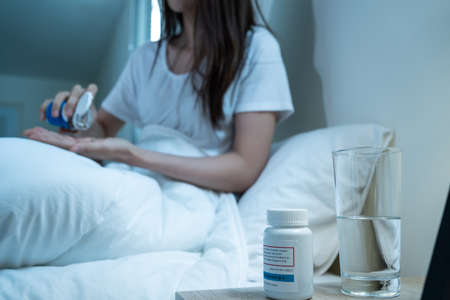 Asian unhappy sick girl in pajamas take medicine before sleep on bed. Attractive beautiful young woman feel frustrated to take many drug to in dark night in bedroom at home. Healthcare-medical conceptの写真素材