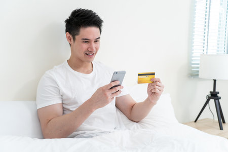 Asian attractive male enjoy shopping online on bed at home in morning. Handsome man sitting in...
