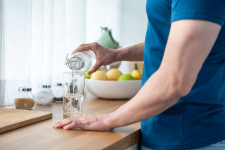 Close up hands of active strong man pouring clean water into glass. Attractive thirsty male drink or take a sip of mineral natural in cup after wake up for health and wellbeing in kitchen in house.の写真素材